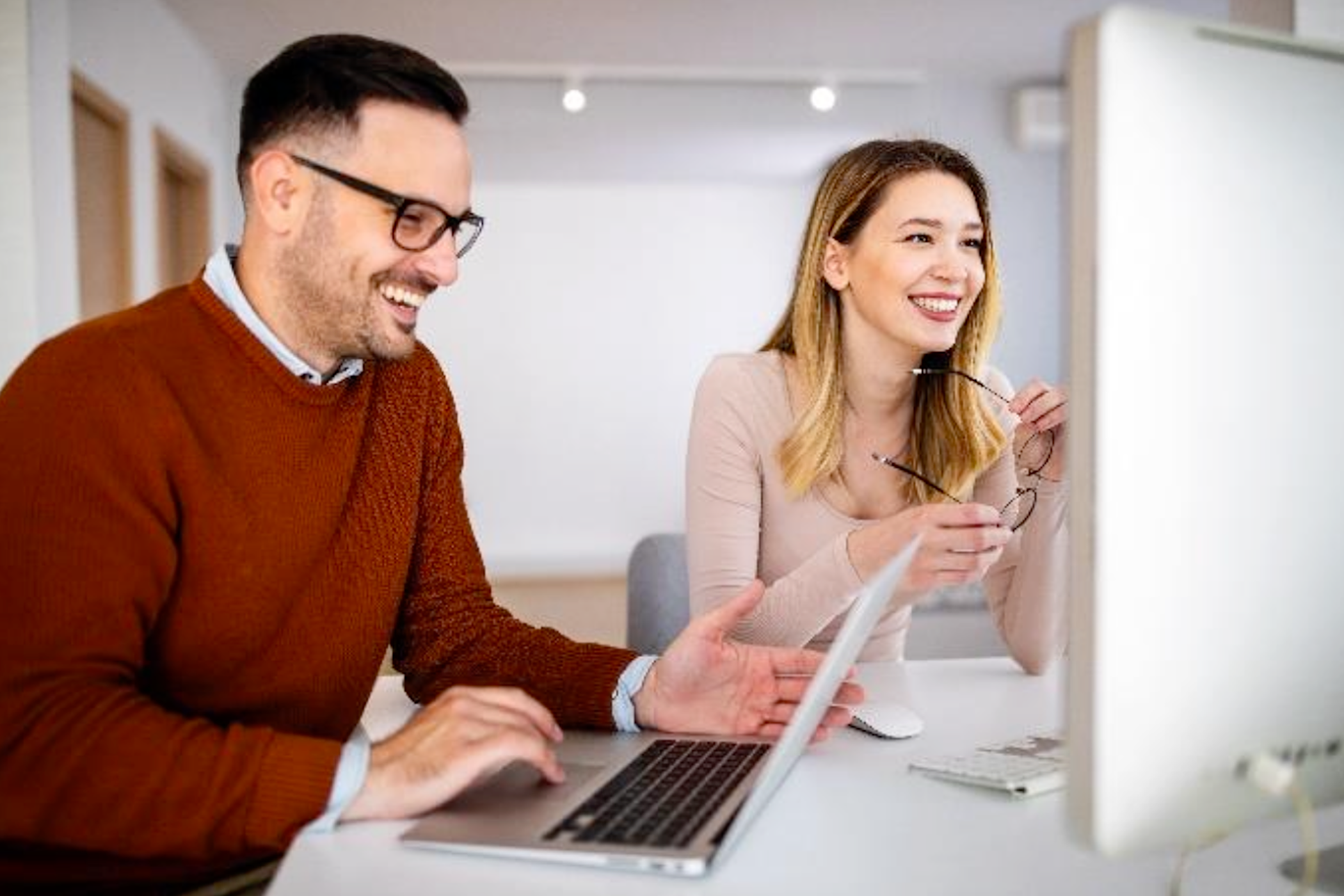 Zwei Mitarbeitende, die am Pc sitzen und lachen. 