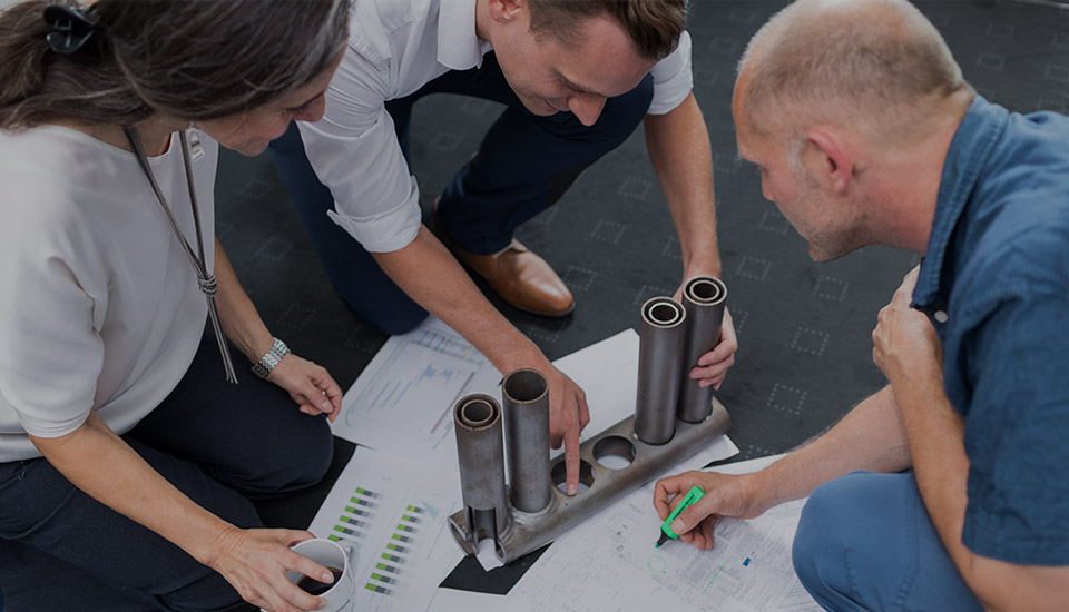 Drei Personen begutachten gemeinsam ein metallisches Bauteil und technische Zeichnungen – Zusammenarbeit in Technik und Vertrieb