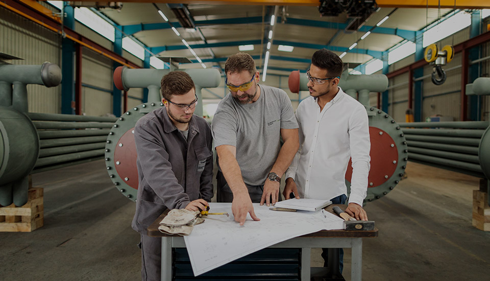 Ein Ausbilder erkl&auml;rt zwei jungen M&auml;nnern technische Zeichnungen in einer Werkshalle &ndash; Ausbildung im industriellen Umfeld