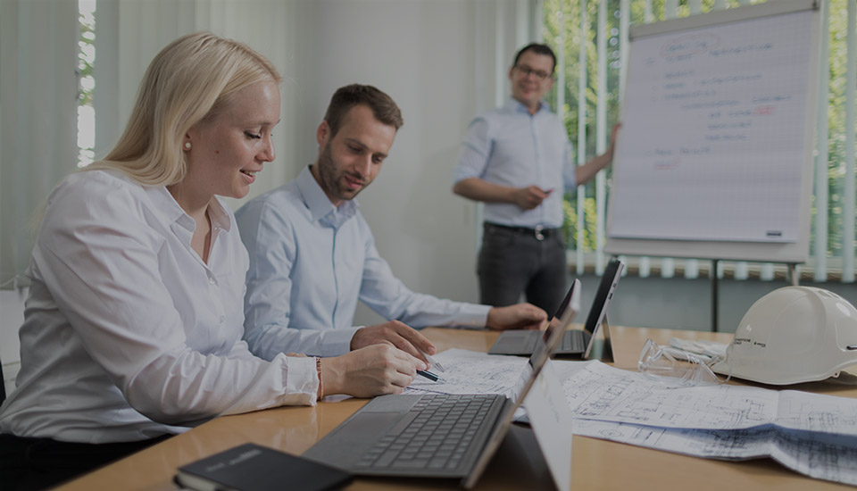 Zwei Mitarbeitende arbeiten gemeinsam an technischen Zeichnungen am Laptop, w&auml;hrend ein Kollege im Hintergrund an einem Flipchart pr&auml;sentiert &ndash; Teammeeting im B&uuml;ro