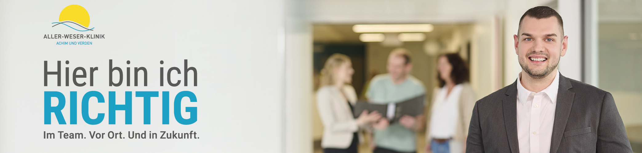 Freundlich l&auml;chelnder Mann in dunklem Anzug und wei&szlig;em Hemd steht im Vordergrund eines Klinikflurs. Im Hintergrund vier Kolleginnen und Kollegen in heller Kleidung im Gespr&auml;ch, eine Person h&auml;lt einen Aktenordner. Links im Bild Logo der Aller-Weser-Klinik mit Slogan &sbquo;Hier bin ich richtig &ndash; Im Team. Vor Ort. Und in Zukunft.