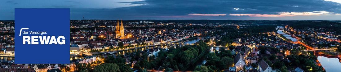 Ein Blick über Regensburg bei Nacht, der beleuchtete Dom und das REWAG-Logo in einem blauen Kasten.