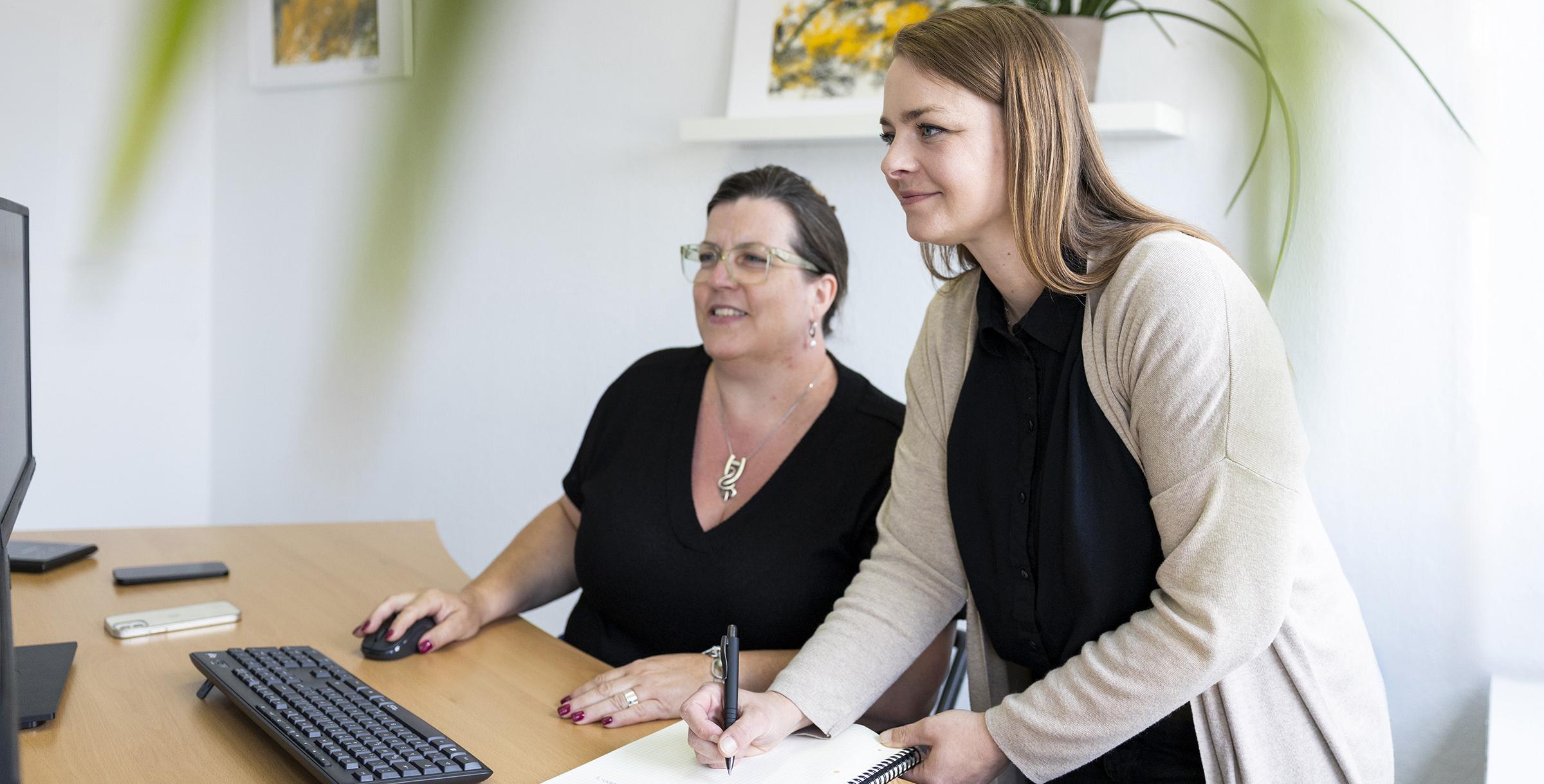 Zwei Frauen an einem Schreibtisch, eine sitzt vor einem Computer mit Tastatur und Maus, die andere steht und schreibt in ein Notizbuch