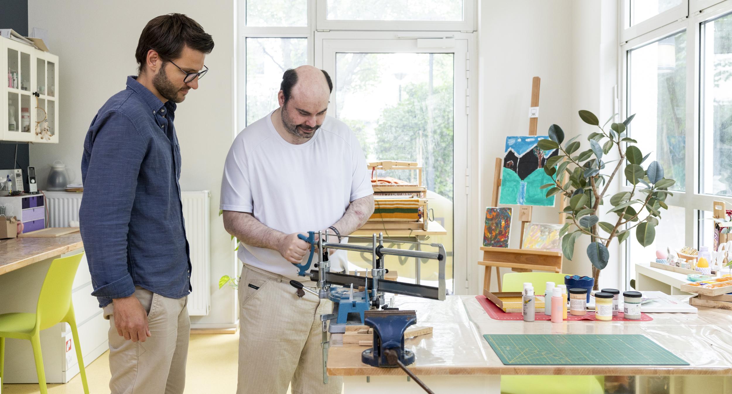 Zwei Männer stehen in einem hellen Atelier mit großen Fenstern neben einem Arbeitstisch, auf dem eine Werkbank mit Schraubstock und verschiedene Malutensilien stehen